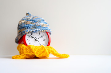 Wall Mural - red round alarm clock in knitted wool blue hat and yellow scarf on a white background. winter time concept. winter season. cozy and warm. analog time 10:10. Space for text