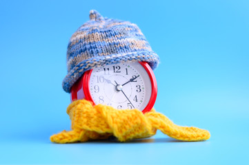 Wall Mural - red round alarm clock in knitted wool blue hat and yellow scarf centered on a blue background. winter time concept. winter season. cozy and warm. analog time 10:10. space for text