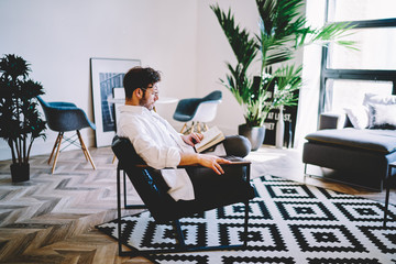 Thoughtful man dressed in casual look resting in comfortable chair and reading interesting bestseller about youth lifestyle, young hipster guy relaxing during literature hobby in home interior