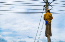 The Technician Is Climbing On The Electric Pole For Repairing Electric Problems In The Background Of Cloudy Blue Sky, Concept Of Risk For Hazardous Work.