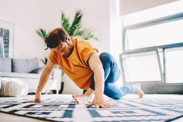 Fototapete - Young man doing stretching exercises during morning energetic workout to develop flexibility and lead a healthy lifestyle in modern flat with stylish interior.Hipster guy practicing sportive training