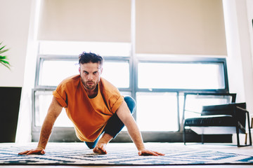 Fototapete - Portrait of concentrated and motivated young man leans with hands on the floor and looking at camera while doing stretching exercises to practice morning yoga at home interior of modern flat