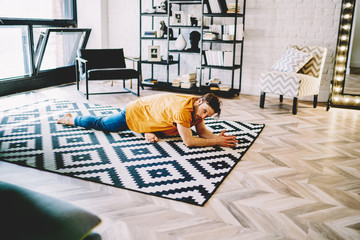 Fototapete - Young man doing stretching exercises on cozy carpet to develop flexibility and maintain vitality in modern flat.Male lover of yoga practicing energetic and healthy lifestyle in apartment
