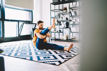 Fototapete - Concentrated young man stretching leng doing yoga exercises during morning workout in modern flat with stylish interior.Sportsman is engaged in physical training on cozy carpet in apartment