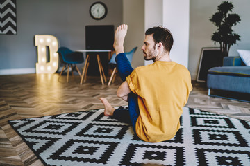 Fototapete - Back view of young man stretching leg sitting on stylish carpet in modern apartment.Hipster guy lover of yoga practicing morning exercises to support vitality and to lead healthy lifestyle