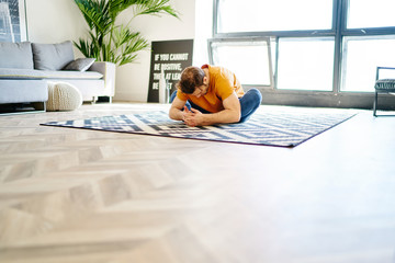 Fototapete - Healthy young man sitting in pose lotus on carpet and doing yoga exercises during morning workout in modern apartment.Hipster guy practicing meditation in stylish flat with home interior