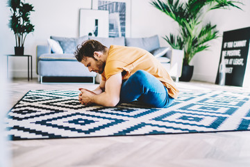 Fototapete - Side view of young man sitting in lotus pose on carpet and doing stretching exercises in modern apartment.Motivated hipster girl practicing yoga and healthy lifestyle at home interior