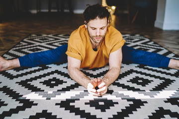 Fototapete - Concentrated young man with closed eyes sitting in twine on carpet and doing stretching exercises during morning training at home interior.Motivated sportsman develop flexibility in apartment