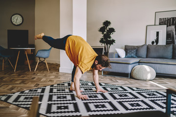 Fototapete - Young man holding leg up during morning yoga exercises on cozy carpet at home apartment with stylish interior.Hipster guy lead healthy lifestyle doing stretching training in stylish flat
