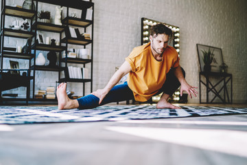 Fototapete - Young man stretching leg doing yoga exercises in the morning at modern home interior.Motivated hipster guy practicing sportive exercises to support body shape in comfortable apartment
