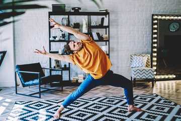 Fototapete - Motivated young man bends and stretching his hands to the side during morning exercises at home interior.Hipster guy doing yoga and lends healthy lifestyle standing on carpet in apartment