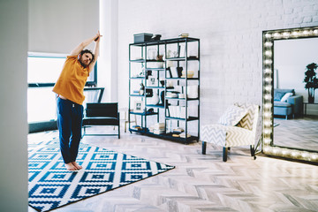 Fototapete - Experienced young man standing on carpet and holding hands in namaste during yoga exercises in home apartment.Hipster guy support healthy viability during morning meditation in flat