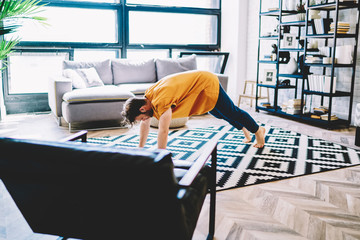 Fototapete - Motivated young man lover of yoga engaged in sport and leads healthy lifestyle at home apartment.Hipster guy practicing sportive poses on carpet to support body shape and vitality in comfortable flat