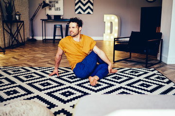 Fototapete - Professional young man doing stretching exercises on comfortable carpet during morning workout in morning time.Experienced sportsman warming up before workout at home interior of apartment