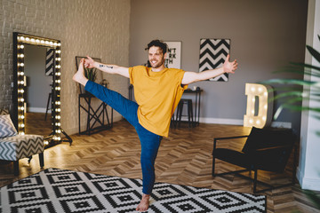 Fototapete - Happy young man holding leg in hand and standing on carpet doing stretching exercises during morning training in home apartment.Positive male lover of yoga practising meditation in different poses