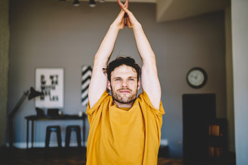 Fototapete - Handsome young man with blue eyes holding hands in namaste above the head above the head during meditation and relaxation in modern apartment.Motivated hipster guy doing yoga exercises during training