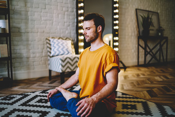 Fototapete - Calm young man with closed eyes sitting in lotus pose on comfortable carpet meditating in morning time at home interior.Hipster guy doing yoga to support flexibility and viability every day
