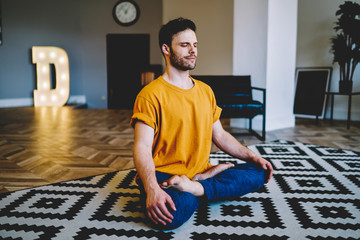 Fototapete - Young man with closed eyes smiling during meditation sitting in lotus pose on cozy carpet in modern apartment.Calm hipster guy 20 years old engaged in yoga in morning time at home interior