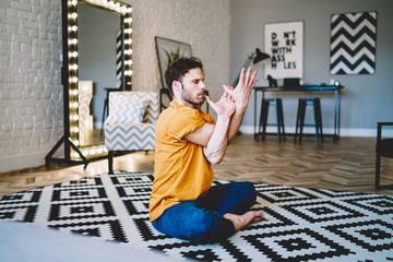 Fototapete - Professional male lover of yoga sitting in lotus pose and stretching hands during morning training at home interior.Calm young man practicing asana during sportive exercises in apartment