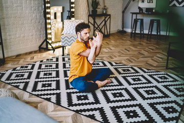 Fototapete - Young man stretching hands during asana pose sitting in lotus on comfortable carpet in modern apartment.Hipster guy 20s old practicing meditation during morning training at home interior