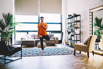 Fototapete - Concentrated young man standing in asana pose practising professional yoga exercises on comfortable carpet in modern apartment.Calm hipster guy standing on one leg enjoying harmony and relaxation