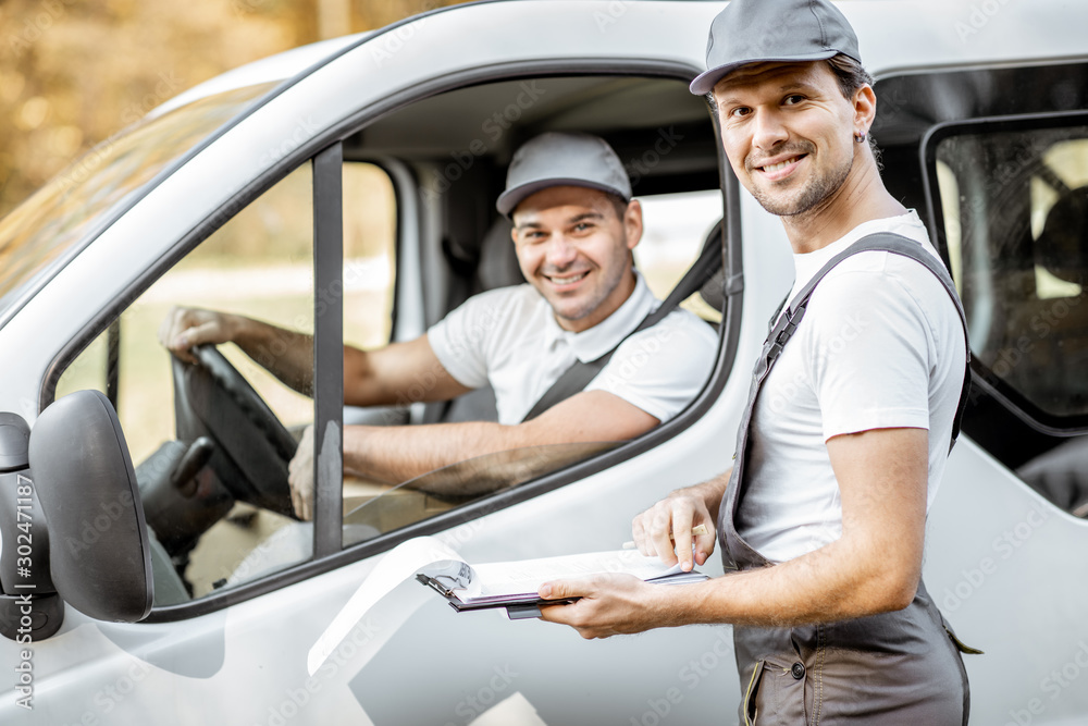 Portrait of a cherfull delivery company employees in uniform delivering goods by cargo van vehicle, mover with a car driver checking delivery list - obrazy, fototapety, plakaty 