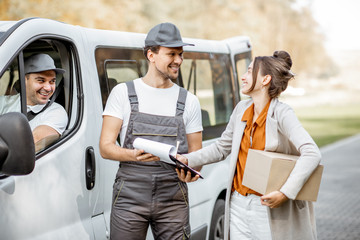 Delivery company employees in uniform delivering goods to a client by cargo van vehicle, woman signing documents and receiving a parcel outdoors