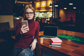 Wall Mural - Attractive female student in spectacles searching information for course work on knowledge websites while sitting near cafeteria table with notebook with mock up screen for your internet advertising