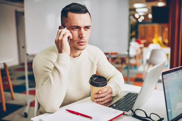 Serious professional worker having mobile phone conversation about business sitting in office, young male entrepreneur checking banking account calling ro service operator during coffee break