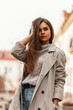 Pretty urban young woman straightens hair. Beautiful stylish girl a fashion model in a fashionable trench coat in a vintage gray knitted sweater walks around the city near the buildings. Spring style.