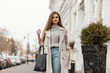 © alones - Pretty trendy young woman in a stylish trench coat in a vintage knitted sweater in blue jeans with a leather bag enjoys a walk along the street. Attractive girl in fashionable clothes in the city.