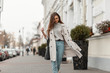 © alones - Stylish model of a young woman in fashionable seasonal clothes with bag walks around the city near a vintage building on a warm autumn day. Beautiful girl in trendy outerwear outdoors.