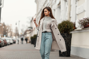 Wall Mural - Model of a young beautiful woman in a fashion trench coat in a stylish knitted sweater with a leather black bag walks on the city near a white building. Elegant pretty trendy girl travels the street.