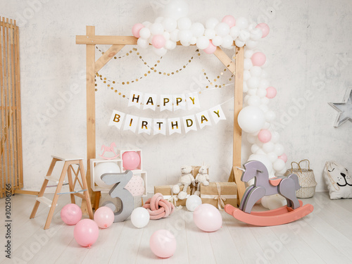 Birthday decorations with wooden arch, gifts, toys, balloons, garland and figure 3 for little baby party on a white wall background.