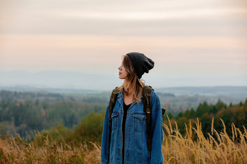 Wall Mural - Style girl in denim jacket and hat with backpack in countryside with mountains on background