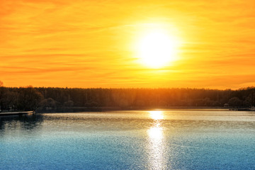 dramatic sunset over pond with sun and outdoor scenery reflecting on lake water surface landscape against orange sky background. Wide view of sunrise with sun above horizon at dawn time wallpaper