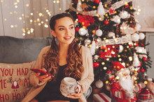 Happy Woman Sitting On The Sofa And Looking At Window And Hold Mobile Phone. Christmas Tree With Decoration Is Next To Her. Holidays And Technology Concepts.