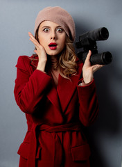 Wall Mural - Young woman in red coat with binoculars on grey background