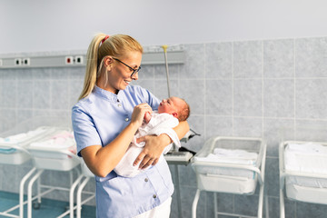 Young nurse standing in maternity ward and holding newborn baby in her arms. After birth concept.