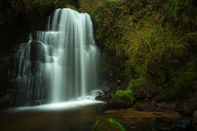 Long Exposure Of Secluded Waterfall, Silky Smooth Water Flowing Down Rocky Face, With Grass Covered Rocks In Foreground, And Surrounded By Green Undergrowth