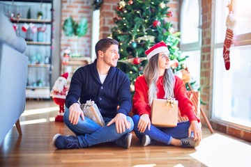 Sticker - Young couple wearing santa claus hat sitting on the floor around christmas tree at home looking to side, relax profile pose with natural face with confident smile.