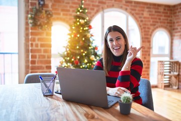 Poster - Beautiful woman sitting at the table working with laptop at home around christmas tree with a big smile on face, pointing with hand and finger to the side looking at the camera.