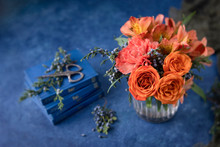 Vase Of Orange Roses, Carnations, And Alstroemeria In Clear Vase On Blue Textured Background; Vintage Scissors On Small Blue Books In Background; Sprigs Of Blue Cedar Berries Surround Vase.