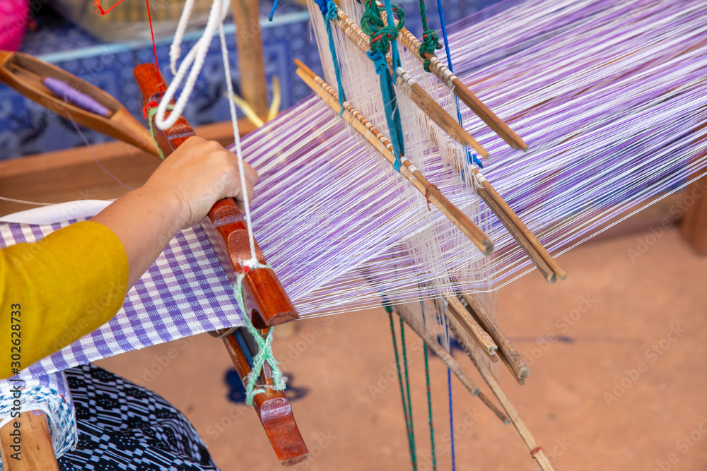 Cotton weaving. Woman hand weaving cotton on manual loom. Thai cotton handmade. Homespun fabric process. The process of fabric weaving in vintage weaver machine. Selective focus. Adobe Stock