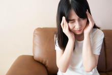Young Asian Woman Suffering From Strong Headache And Sitting On Couch At Home. Cause Of Headache Include Migraine, Tension Headache, Stress, Brain Tumor Or Stroke. Health Care And Medical Concept.