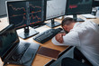 © standret - Tired businessman sleeping on the workplace by leaning on the table with multiple screens on it. Stock information on displays