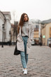 © alones - Attractive brown-haired young woman in fashionable spring clothes in white stylish sneakers with a leather bag walks along the street near vintage buildings.Cute girl walks on a stone road in the city