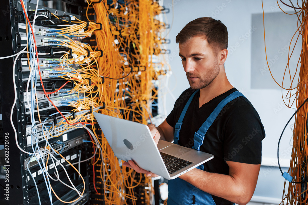 Young man in uniform and with laptop works with internet equipment and wires in server room - obrazy, fototapety, plakaty 