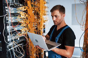 Wall Mural - Young man in uniform and with laptop works with internet equipment and wires in server room