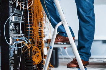 Wall Mural - Man in uniform stands on the ladder and works with internet equipment and wires in server room